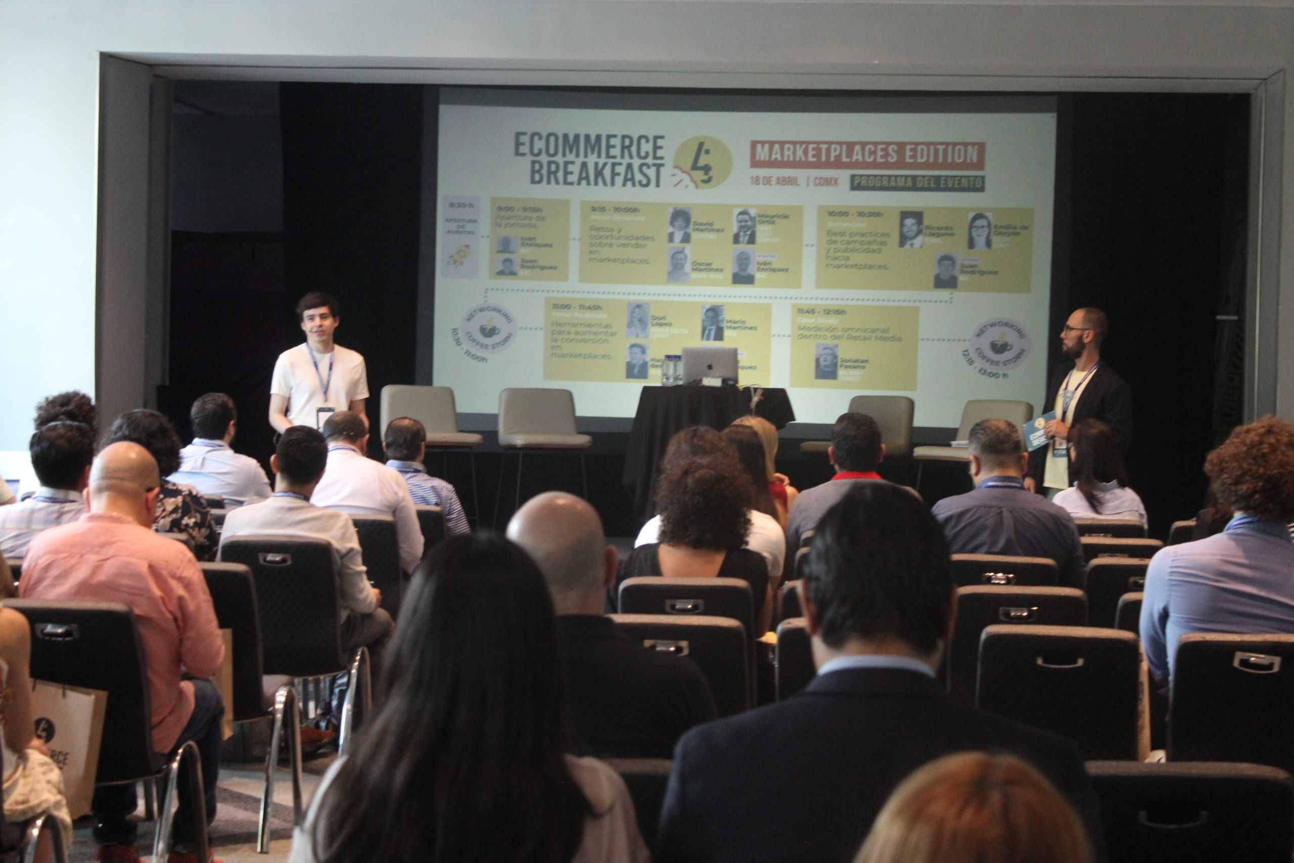 Pantalla de proyección que muestra una diapositiva del evento. Dos presentadores se encuentran en los extremos del escenario y el público está sentado frente a ellos en una sala de conferencias.

Texto transcrito visible: "ECOMMERCE BREAKFAST" "MARKETPLACES EDITION" "18 DE ABRIL | COMEX" "PROGRAMA DEL EVENTO"