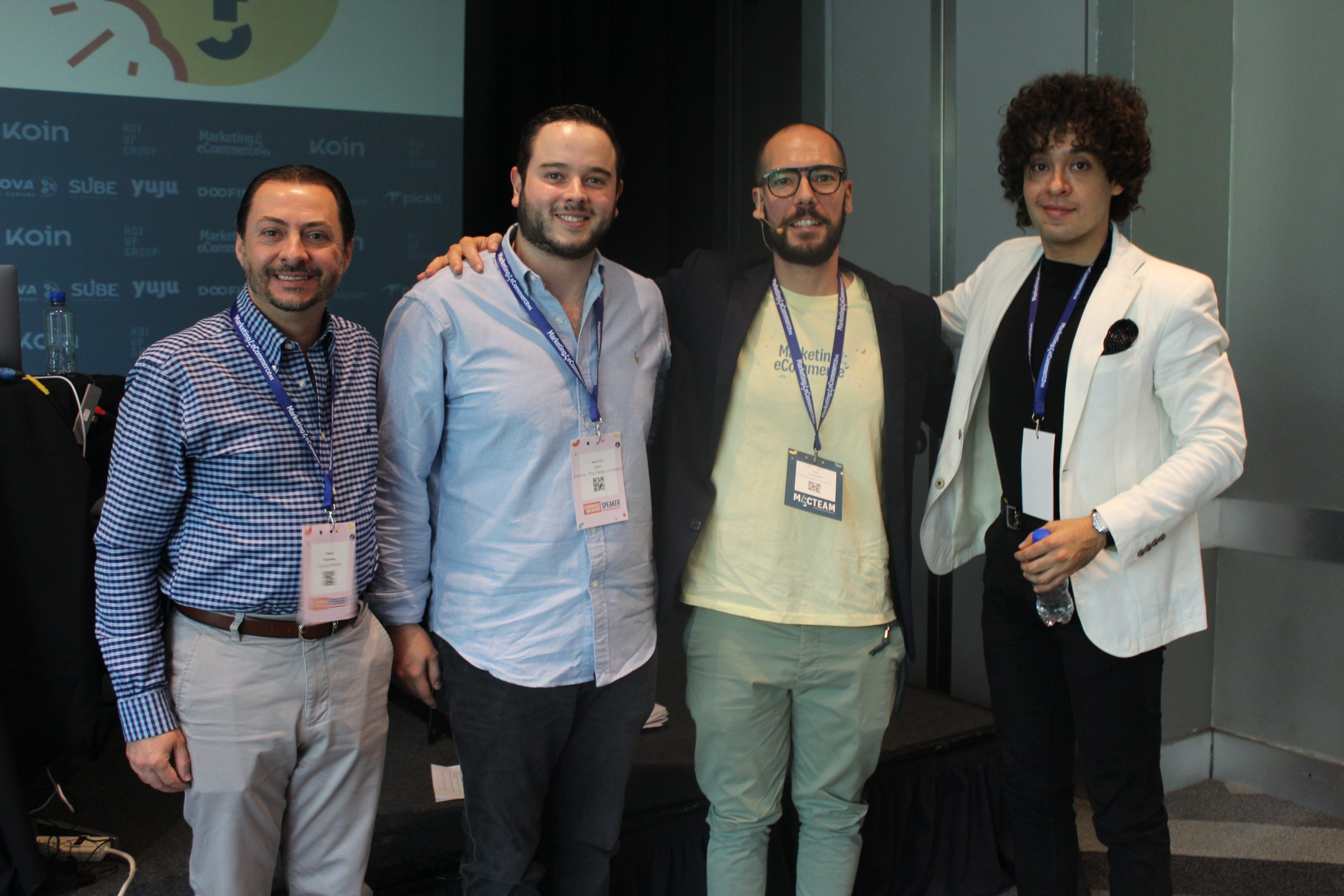 Cuatro hombres, de pie muy cerca unos de otros, sonriendo y posando abrazados en un pequeño escenario de conferencias, llevan cordones y credenciales, con vestimenta informal a formal. Texto visible: "koin" repetido en el fondo.