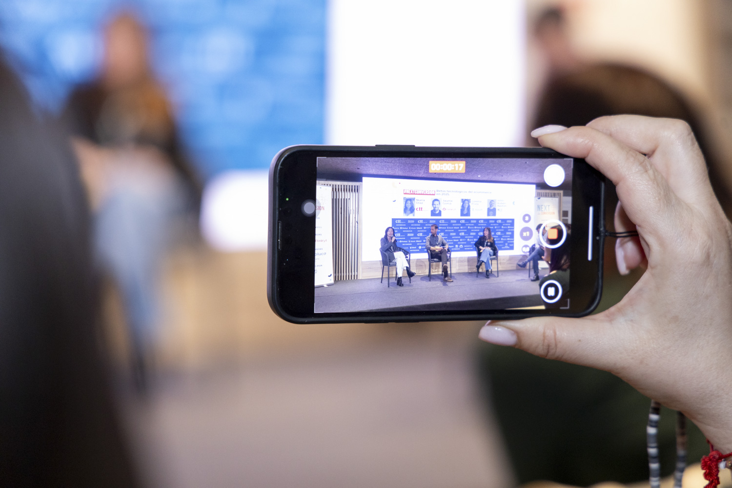 Un teléfono inteligente (objeto) graba a cuatro oradores (acción) en un escenario de conferencias, mientras que la escena real fuera del teléfono aparece borrosa (contexto).  Texto visible en el teléfono: 00:00:17