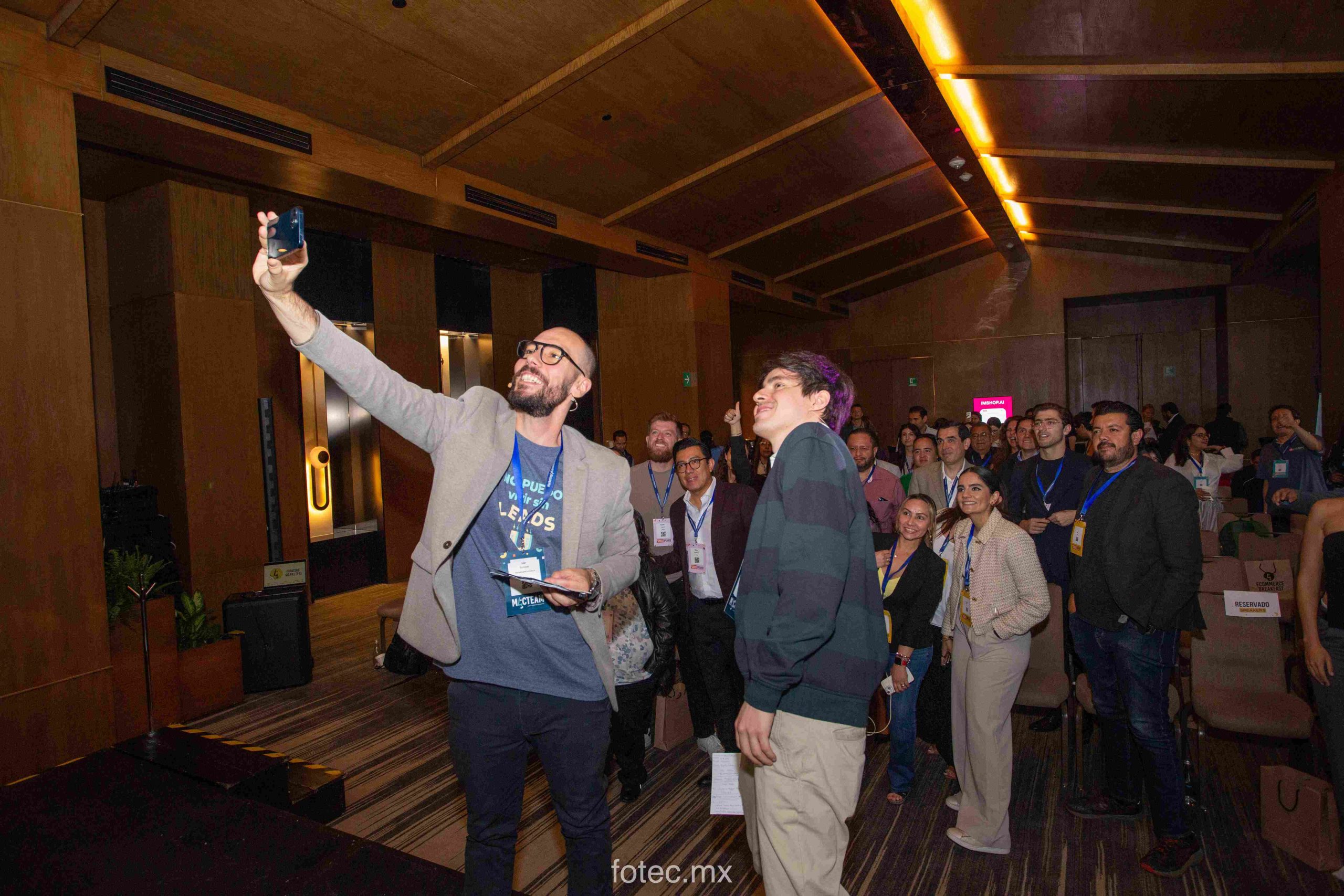 Un hombre con un teléfono inteligente se toma una selfie con una multitud sonriente en una sala de conferencias con paneles de madera. Texto: "fotec.mx" (abajo), "RESERVADO" (letrero en la silla), "IMSHOPAI" (letrero rosa), "LEADS" (en una camiseta).