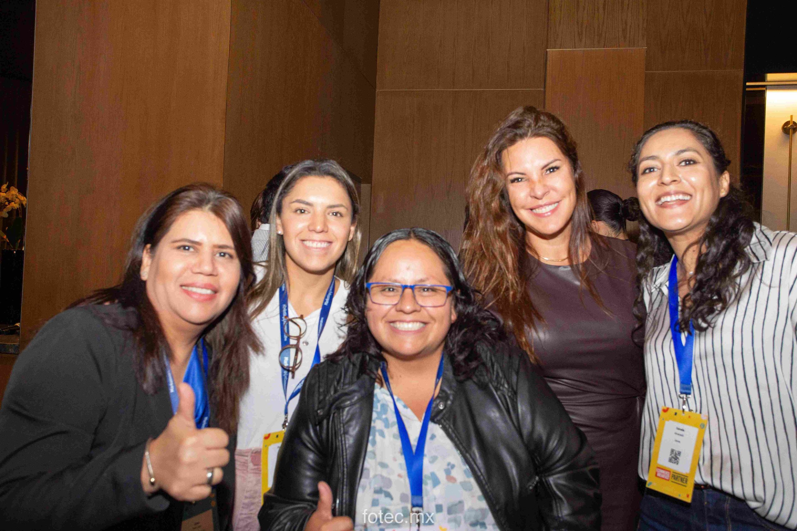 Grupo de cinco mujeres sonriendo y levantando el pulgar, posando juntas en un entorno de conferencia con cordones y credenciales contra paneles de madera. Texto visible: "fotec.mx", "PARTNER".