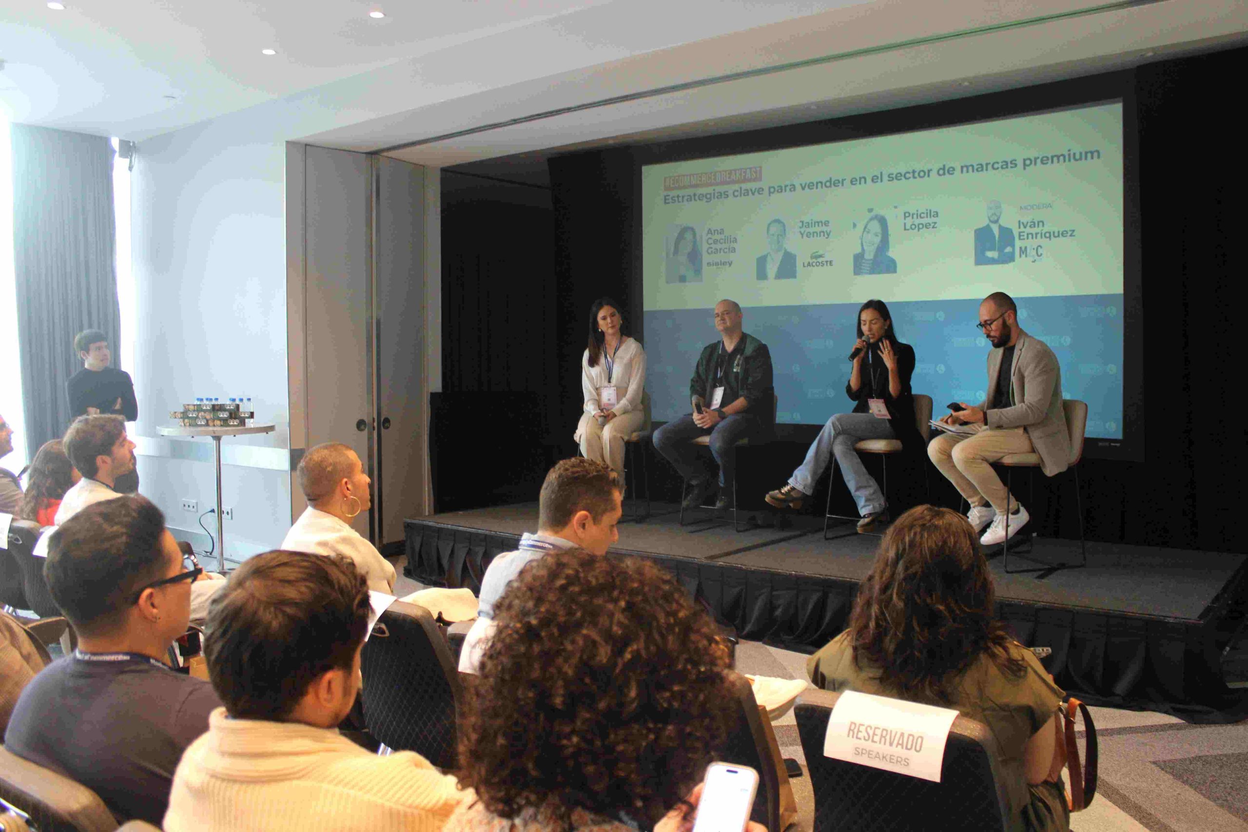 Panel de cuatro oradores sentados en un pequeño escenario discutiendo ideas mientras una audiencia escucha en una sala de conferencias. La diapositiva dice: "Conversación clave para vender en el sector de marcas premium". Un letrero en una silla dice "RESERVADO".