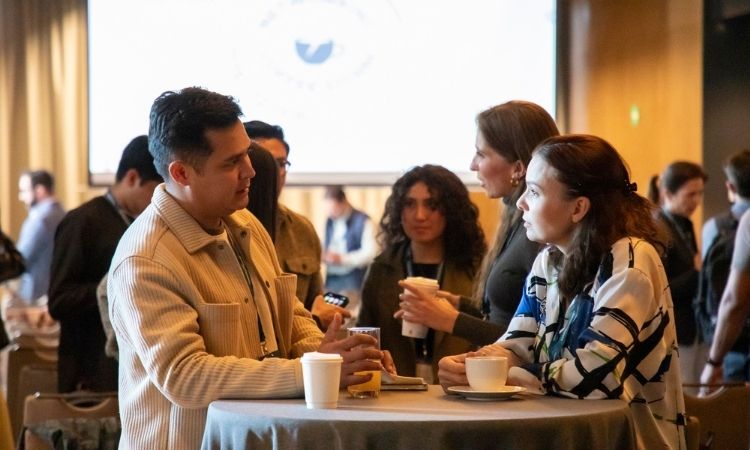 Tres adultos conversan alrededor de una mesa redonda alta, sosteniendo tazas de café y un teléfono; otros asistentes se mezclan en una sala de conferencias con poca luz y una pantalla de proyector al fondo.