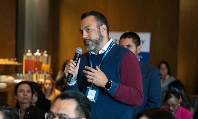 Un hombre, con un micrófono en la mano, formula una pregunta mientras permanece de pie entre los asistentes sentados a una conferencia en una sala de reuniones con poca luz.