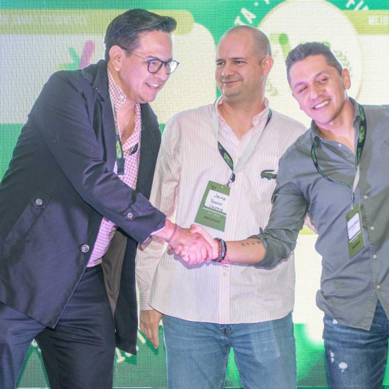 2023 premiosecom Tres hombres en un escenario: dos se dan la mano mientras el tercero observa y sonríe, todos con credenciales y cordones, sobre un fondo iluminado en verde. Texto en la credencial central: "Jaime Yamir Lacote".