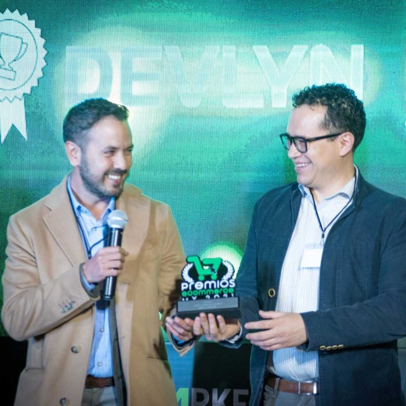 22premiosecommercemx Un trofeo es entregado y sostenido por dos hombres sonrientes en el escenario; uno habla por un micrófono frente a un fondo LED verde. Texto en la imagen: "premios e-commerce", "DEVLYN".