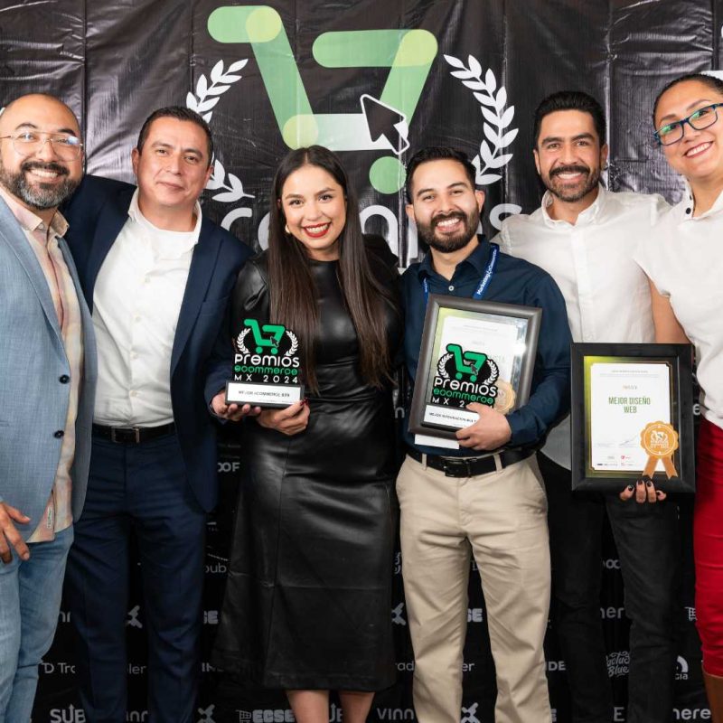24premiosecommercemx Objeto: Seis adultos sosteniendo trofeos y un certificado enmarcado. Acción: Sonriendo y posando para una foto de celebración. Contexto: Evento en interiores frente a un fondo negro con la marca y un logotipo verde. Texto visible: "PREMIOS".