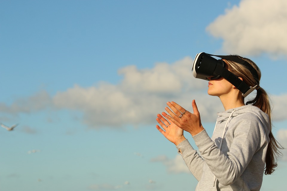 Una persona que lleva puesto un casco de realidad virtual extiende las manos hacia adelante como si interactuara o aplaudiera, de pie al aire libre bajo un cielo azul brillante con nubes dispersas y gaviotas a lo lejos.