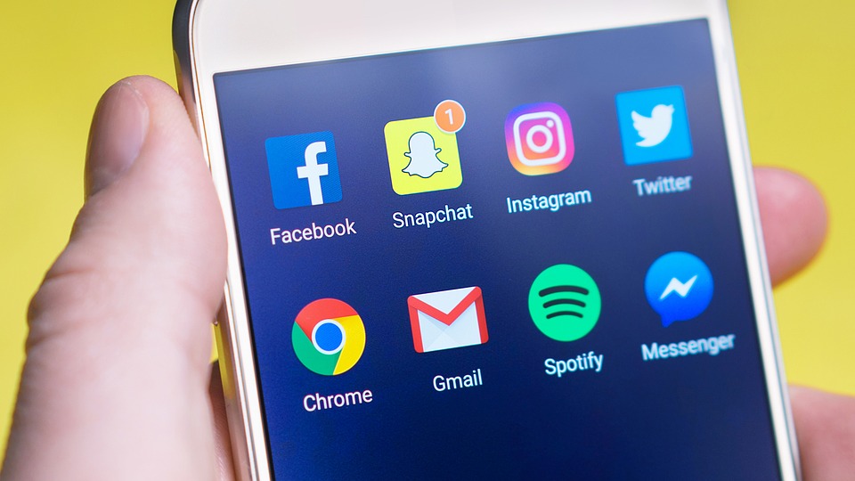 Pantalla de smartphone con aplicaciones de redes sociales y utilidades, sostenida por una mano sobre un fondo amarillo; Snapchat muestra una notificación con el número "1". Texto: Facebook, Snapchat, Instagram, Twitter, Chrome, Gmail, Spotify, Messenger.