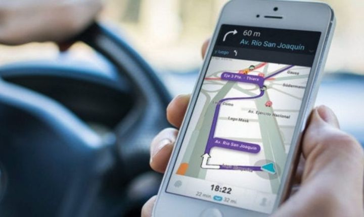 Smartphone mostrando una aplicación de navegación (objeto) con una ruta morada (acción) sostenida por una mano dentro de un coche cerca del volante (contexto). Texto en pantalla: "60 m", "Av. Río San Joaquín", "18:22".