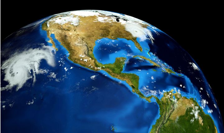 El planeta Tierra gira en el espacio, mostrando América del Norte y Central, el Caribe y América del Sur; formaciones de nubes se desplazan sobre océanos azules y masas terrestres de color verde parduzco que contrastan con un fondo negro.