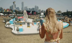 Una mujer sostiene un teléfono mientras observa un modelo en miniatura del mapa del festival con los puestos y escenarios etiquetados: MILLER LITE, MILLER LITE STAGE, HONDA, BMI, BARTON SPRINGS, TITO'S, T-MOBILE, AMERICAN EXPRESS, EXIT.