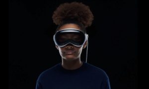 Persona joven con grandes gafas de realidad virtual, de pie y mirando al frente, con los auriculares visibles; el fondo oscuro aísla la parte superior de su cuerpo y su cabello en un retrato de estudio con iluminación suave.