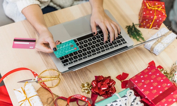 Un teclado de portátil y una mano sosteniendo una tarjeta de crédito —escribiendo y preparándose para pagar— rodeados de regalos envueltos, cintas, lazos y adornos navideños sobre una mesa de madera.