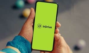El teléfono inteligente muestra una pantalla verde con el logotipo "iD" y la palabra "inDrive"; sostenido con ambas manos, con la manga de una chaqueta azul, sobre un fondo exterior borroso con pelotas de colores.
