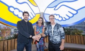 Tres adultos están de pie uno al lado del otro, con las manos entrelazadas frente a ellos; un gran mural colorido de dos manos entrelazadas se alza imponente detrás de ellos en una terraza en la azotea con vistas a la ciudad.