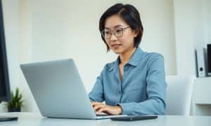 Una joven con gafas teclea en un portátil plateado sobre un escritorio blanco, con un smartphone cerca. El fondo de la oficina es minimalista y luminoso, con una pequeña planta y estanterías.