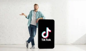Un hombre vestido de manera informal se apoya en un teléfono inteligente de gran tamaño que muestra el logotipo de TikTok y el texto "TikTok", de pie contra una pared de ladrillo blanco en un estudio luminoso y minimalista.
