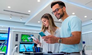 Dos clientes sostienen y prueban teléfonos inteligentes mientras sonríen y comparan características en una tienda de electrónica moderna y luminosa, con televisores de exhibición y estaciones de demostración al fondo.